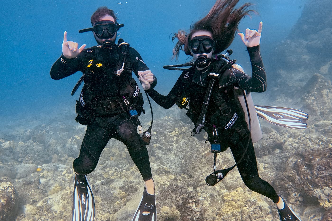 Riley scuba diving with a friend over a reef, both giving the shaka sign
